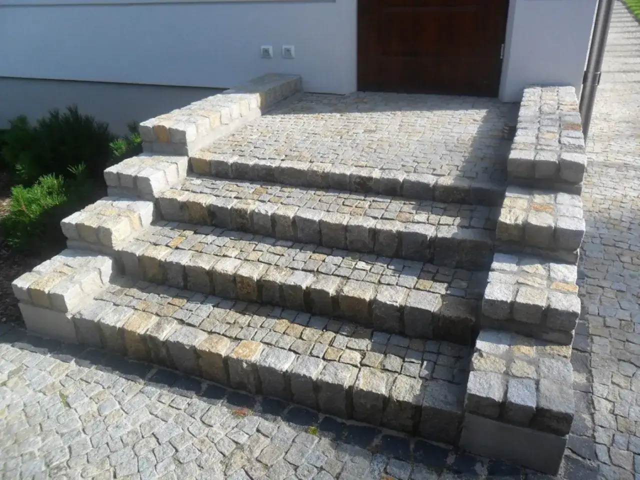 Solidne schody z kostki granitowej prowadzące do drzwi. Wok&oacute;ł zielony krzew i podjazd z tej samej kostki.