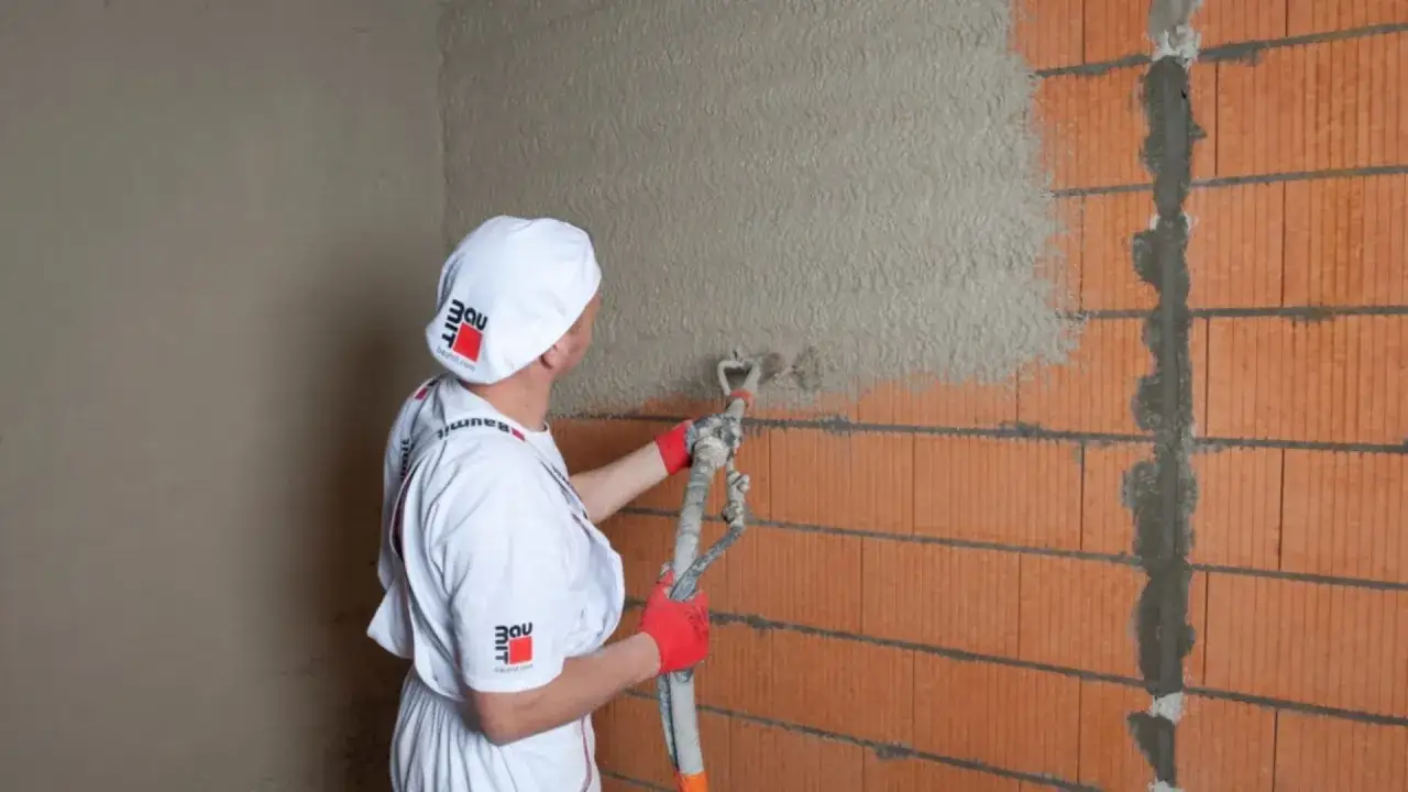 Pracownik nakłada tynki cementowo wapienne na ścianę z pustaków ceramicznych za pomocą agregatu tynkarskiego.