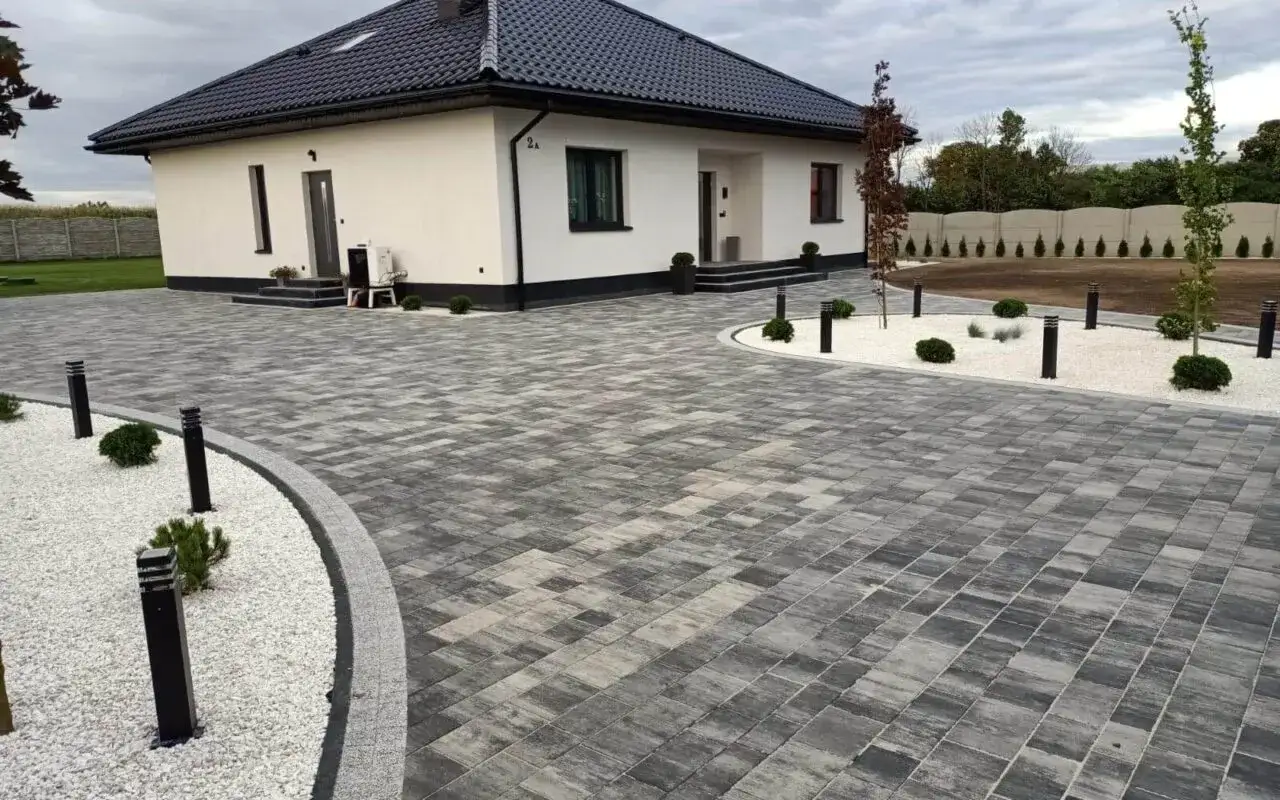 Nowoczesny podjazd z kostki brukowej, kt&oacute;ra ma fazę, tworzy elegancką całość z białym żwirem i minimalistycznym oświetleniem.